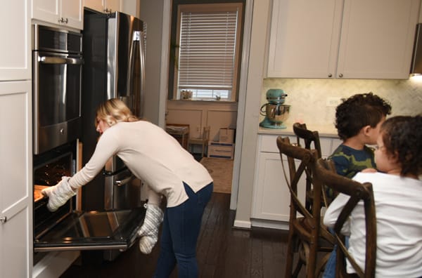 woman taking pan out of oven with children seated behind her