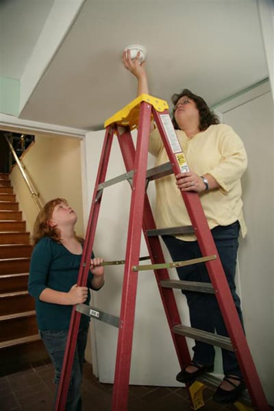 girl holding a ladder for a woman installing a smoke alarm