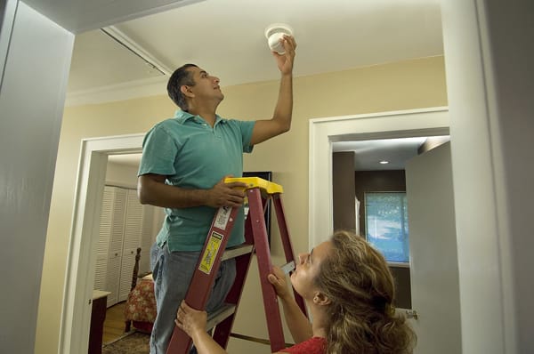 woman holding a ladder for a man installing a smoke alarm