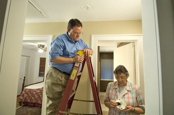 man on a ladder watching an older woman change a smoke alarm battery