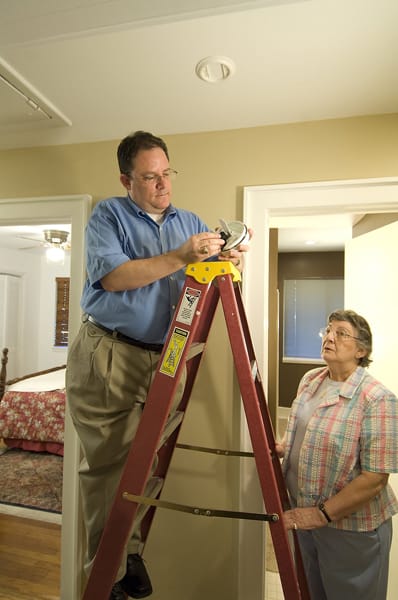 man on a ladder changing smoke alarm battery while older woman watches