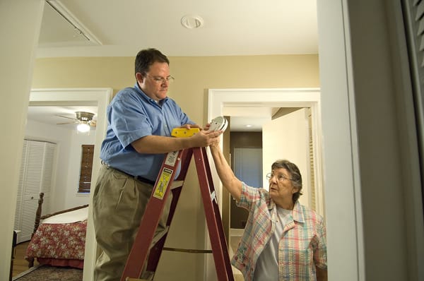 older woman handing smoke alarm to man on a ladder after changing battery