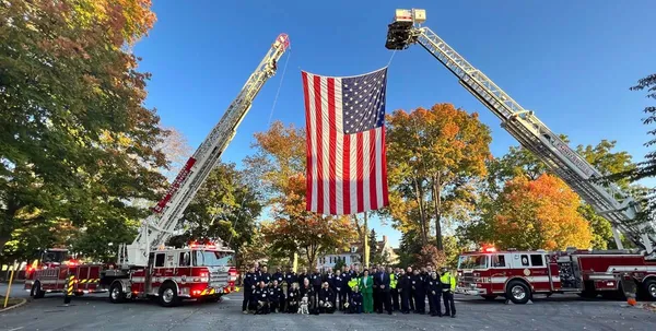 U.S. Fire Administrator Dr. Lori Moore-Merrell and local fire service members are photographed under an American flag raised by 2 ladder trucks.